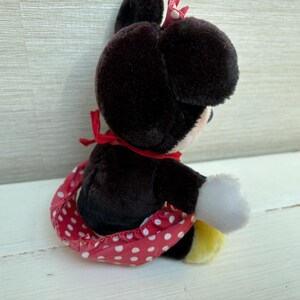 K&ouml;nnte beinhalten: Eine Pl&uuml;schpuppe von Minnie Mouse, mit schwarzem K&ouml;rper, gro&szlig;en Ohren und einem rot-wei&szlig; gepunkteten Rock. Die Puppe hat wei&szlig;e Handschuhe, eine rote Schleife und gelbe Schuhe. Das Spielzeug sitzt auf einer wei&szlig;en Oberfl&auml;che.