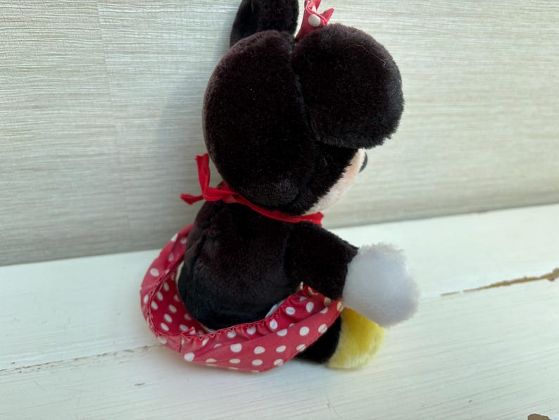 K&ouml;nnte beinhalten: Eine Pl&uuml;schpuppe von Minnie Mouse, mit schwarzem K&ouml;rper, gro&szlig;en Ohren und einem rot-wei&szlig; gepunkteten Rock. Die Puppe hat wei&szlig;e Handschuhe, eine rote Schleife und gelbe Schuhe. Das Spielzeug sitzt auf einer wei&szlig;en Oberfl&auml;che.
