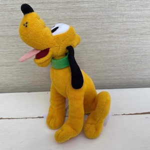 Puede incluir: Un peluche de Pluto, un perro amarillo con orejas negras, una nariz negra y una lengua rosa que sobresale. Tiene un collar verde y ojos blancos. El juguete está hecho de material suave y está sentado erguido.
