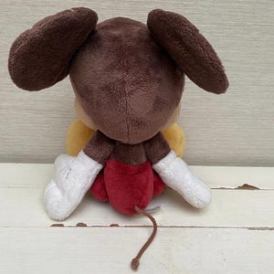 May include: A plush toy of a cartoon character, seen from behind. It has large brown ears, a brown head, and a yellow body. The toy wears red shorts, white gloves, and has a brown tail. It is on a white wooden surface.