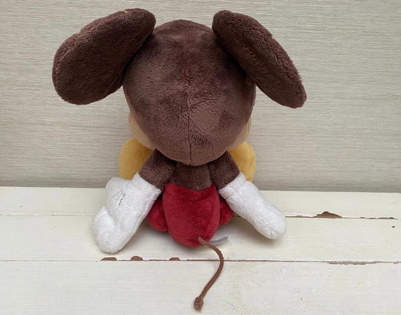 May include: A plush toy of a cartoon character, seen from behind. It has large brown ears, a brown head, and a yellow body. The toy wears red shorts, white gloves, and has a brown tail. It is on a white wooden surface.
