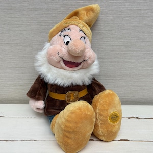 Op de afbeelding: Een knuffel van het personage Happy uit Sneeuwwitje en de zeven dwergen, met een beige muts waarop het woord HAPPY is geborduurd, een witte baard van imitatiebont, een bruine tuniek met een gele riem en grote beige schoenen met een gouden Disney-label.