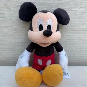 K&ouml;nnte beinhalten: Eine Pl&uuml;schpuppe von Mickey Mouse mit schwarzen Ohren, einem schwarzen K&ouml;rper und einem beigen Gesicht. Die Puppe hat wei&szlig;e Handschuhe, rote Shorts und gelbe F&uuml;&szlig;e. Ein klassischer Disney-Charakter.