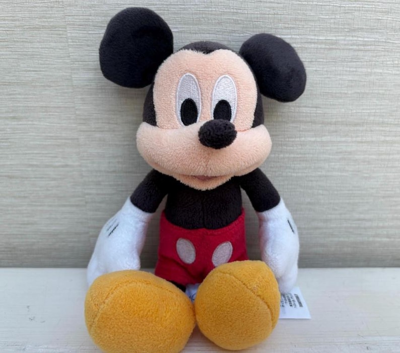 K&ouml;nnte beinhalten: Eine Pl&uuml;schpuppe von Mickey Mouse mit schwarzen Ohren, einem schwarzen K&ouml;rper und einem beigen Gesicht. Die Puppe hat wei&szlig;e Handschuhe, rote Shorts und gelbe F&uuml;&szlig;e. Ein klassischer Disney-Charakter.