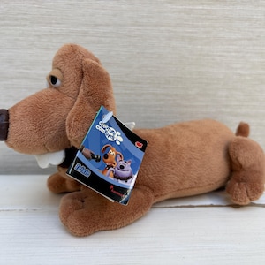 Könnte beinhalten: Ein braunes Plüschhund-Spielzeug mit langen Ohren, einer dunkelbraunen Nase und markanten weißen Zähnen. Am Hals befindet sich ein blau-schwarzes Etikett mit dem Text Creature Comforts und einer Abbildung von zwei Hunden. Das Spielzeug hat einen langen Körper.