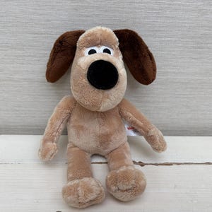 May include: A plush dog toy with light tan fur, dark brown floppy ears, and a large black nose. The character has wide white eyes with black pupils and a small white tag attached to its side. The stuffed animal is in a seated position.