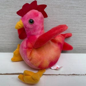 May include: A plush rooster toy in shades of pink, red, and orange. The rooster has a red comb and wattle, yellow beak and feet, and a round black eye. The toy is made of soft fabric and is designed to sit upright.