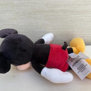 K&ouml;nnte beinhalten: Ein Stofftier von Mickey Mouse, das auf der Seite liegt. Das Spielzeug hat einen schwarzen Kopf und Ohren, ein rosa Gesicht, rote Shorts, wei&szlig;e Handschuhe und F&uuml;&szlig;e sowie gelbe Schuhe. Ein wei&szlig;es Etikett ist am Spielzeug befestigt.