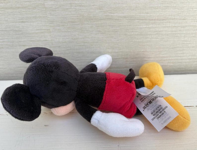 K&ouml;nnte beinhalten: Ein Stofftier von Mickey Mouse, das auf der Seite liegt. Das Spielzeug hat einen schwarzen Kopf und Ohren, ein rosa Gesicht, rote Shorts, wei&szlig;e Handschuhe und F&uuml;&szlig;e sowie gelbe Schuhe. Ein wei&szlig;es Etikett ist am Spielzeug befestigt.