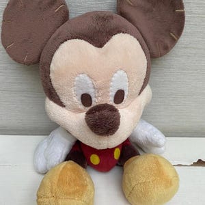 May include: A soft Mickey Mouse toy with brown ears, a brown head, and a cream-coloured face. The toy has a red shirt with yellow buttons, white gloves, and yellow feet. The toy is designed to be soft and cuddly.