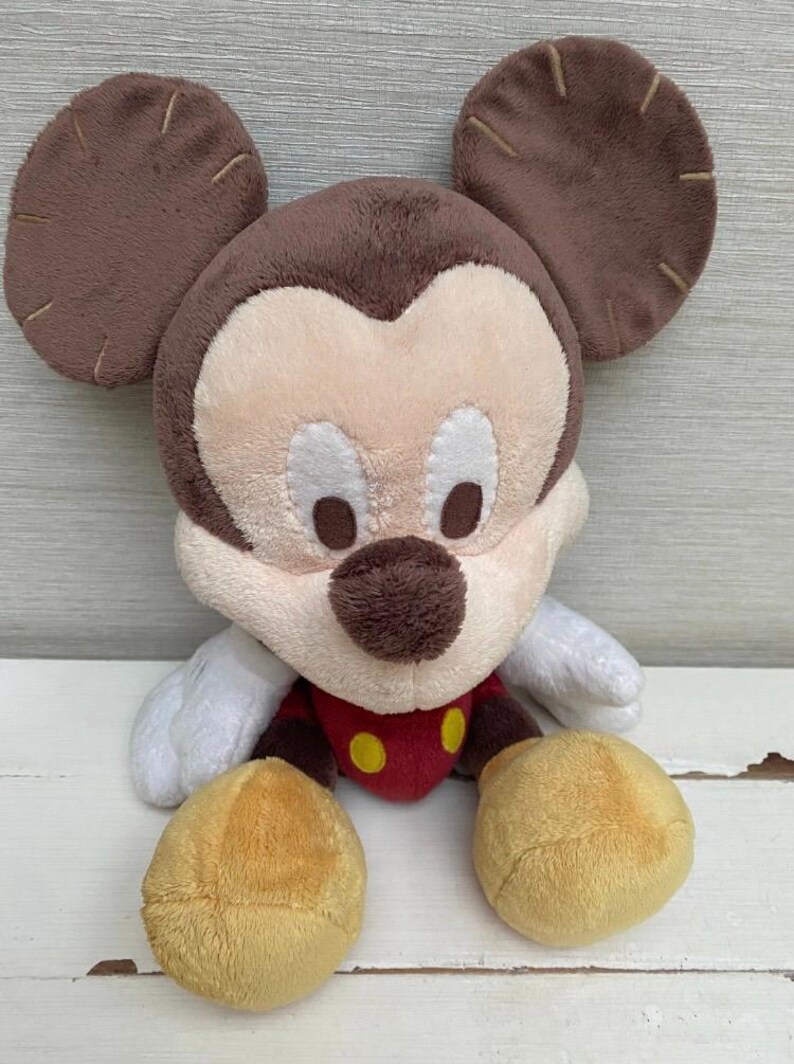 May include: A soft Mickey Mouse toy with brown ears, a brown head, and a cream-coloured face. The toy has a red shirt with yellow buttons, white gloves, and yellow feet. The toy is designed to be soft and cuddly.