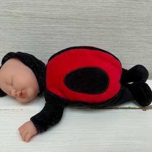 May include: A soft baby doll in a ladybug costume. The doll has a fair complexion, closed eyes, and a black hood with small antennae. The ladybug outfit is red with a black oval on the back and black arms and legs.