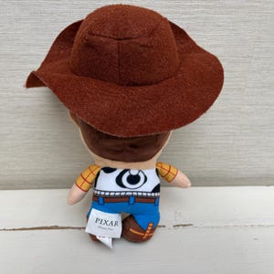 May include: A plush toy of the character Woody from the Pixar movie Toy Story. The toy has a brown cowboy hat, a white shirt with a cow print, blue pants, and brown boots. The Pixar logo is visible on a tag.
