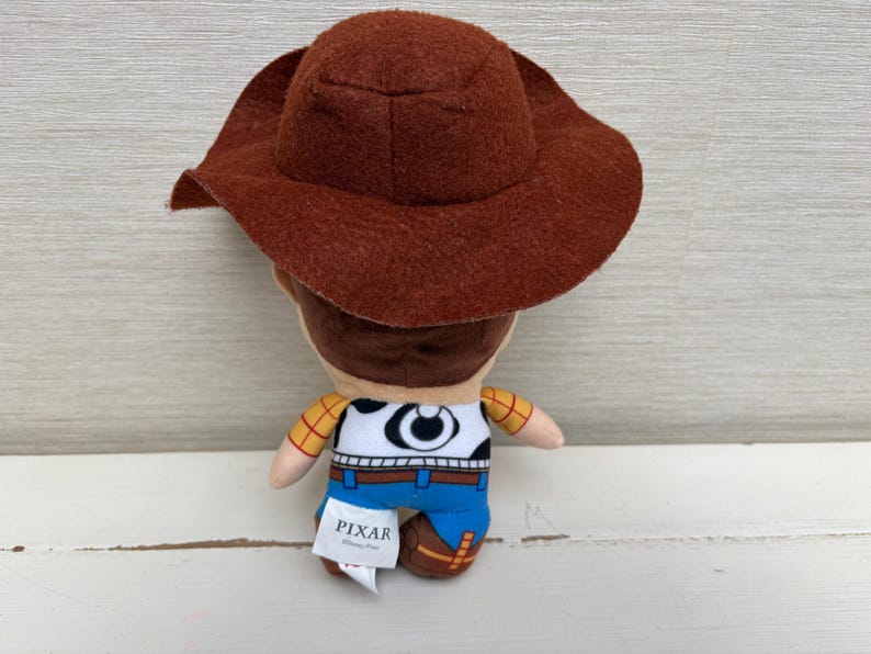 May include: A plush toy of the character Woody from the Pixar movie Toy Story. The toy has a brown cowboy hat, a white shirt with a cow print, blue pants, and brown boots. The Pixar logo is visible on a tag.