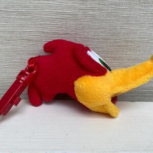 May include: A plush red bird with a yellow beak and a red clip. The bird has white and green eyes. The clip is attached to the bird's body. The bird is lying on a white surface.