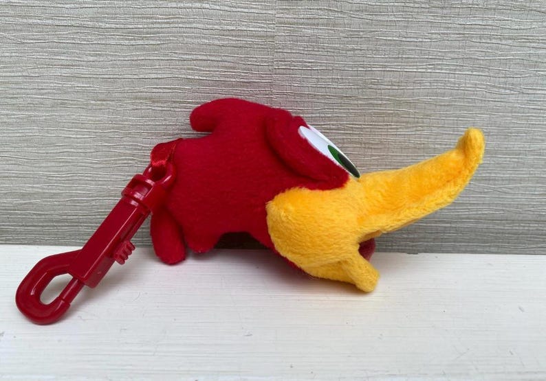 May include: A plush red bird with a yellow beak and a red clip. The bird has white and green eyes. The clip is attached to the bird's body. The bird is lying on a white surface.