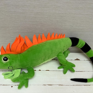 Könnte beinhalten: Ein Leguan-Stofftier mit einem leuchtend grünen Körper, einer Reihe leuchtend orangefarbener Filzstacheln auf dem Rücken und einem langen Schwanz mit abwechselnd schwarzen und grünen Streifen. Das Plüschtier hat große dunkle Augen und einen hellgrünen Kehlsack.