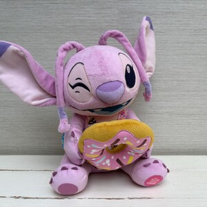 Puede incluir: Un peluche de color lavanda con orejas grandes y una expresi&oacute;n gui&ntilde;adora, sosteniendo una rosquilla amarilla y rosa. El peluche tiene una boca azul y est&aacute; sentado sobre una superficie blanca.