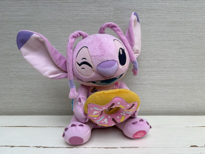 Puede incluir: Un peluche de color lavanda con orejas grandes y una expresi&oacute;n gui&ntilde;adora, sosteniendo una rosquilla amarilla y rosa. El peluche tiene una boca azul y est&aacute; sentado sobre una superficie blanca.