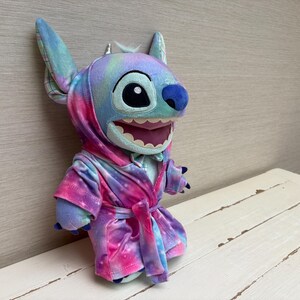 May include: A plush Stitch doll wearing a tie-dye robe in shades of blue, purple, and pink. The doll has large ears, a blue nose, and a wide, toothy grin. The robe is tied at the waist with a matching belt.