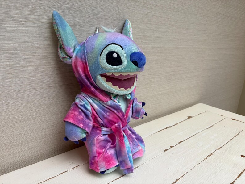 May include: A plush Stitch doll wearing a tie-dye robe in shades of blue, purple, and pink. The doll has large ears, a blue nose, and a wide, toothy grin. The robe is tied at the waist with a matching belt.