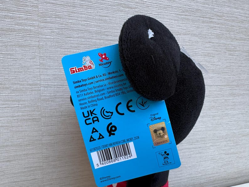May include: A black plush Mickey Mouse toy with a blue tag. The tag features the Simba and Nicotoy logos, along with text and a barcode. The toy is a Disney product.