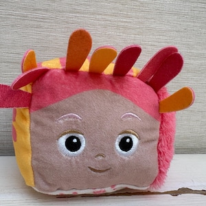 Puede incluir: Un juguete de peluche en forma de cubo con un rostro bordado que incluye ojos, cejas y una pequeña sonrisa. El cubo está cubierto de tela suave rosa y amarilla, con varias protuberancias de fieltro en forma de pétalos naranjas y rosas.