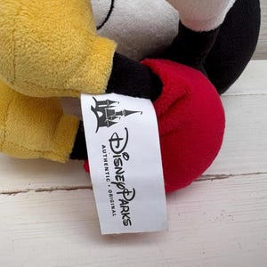 Op de afbeelding: Een Mickey Mouse knuffel met gele handschoenen, een rode torso en een zwarte broek. Het speelgoed heeft een wit gezicht en zwarte oren. Een wit label met het Disney Parks-logo is bevestigd.