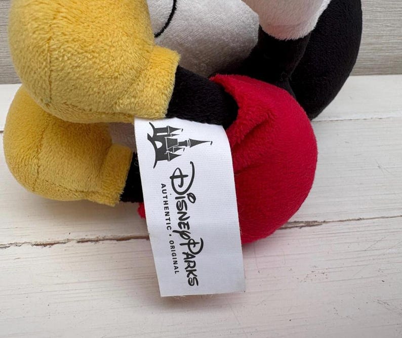 Op de afbeelding: Een Mickey Mouse knuffel met gele handschoenen, een rode torso en een zwarte broek. Het speelgoed heeft een wit gezicht en zwarte oren. Een wit label met het Disney Parks-logo is bevestigd.