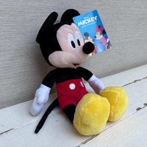 May include: A plush Mickey Mouse doll with black ears, a black tail, and a red shorts with white buttons. The doll has white gloves and yellow shoes. A tag that says "Mickey and Friends" is attached.
