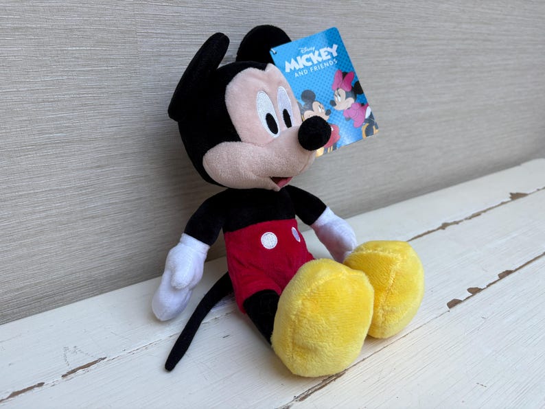 May include: A plush Mickey Mouse doll with black ears, a black tail, and a red shorts with white buttons. The doll has white gloves and yellow shoes. A tag that says "Mickey and Friends" is attached.