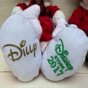 May include: Close-up of a plush doll's white foot coverings, one with "Dilly" in gold script and the other with "Disney Store 2017" in green. The doll's red and white outfit is visible in the background.