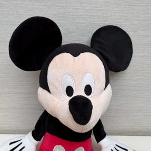 May include: A plush Mickey Mouse toy with large black ears, a black nose, and white gloves. The toy has a red pair of shorts and a cream-colored face. The toy is a classic Disney character.