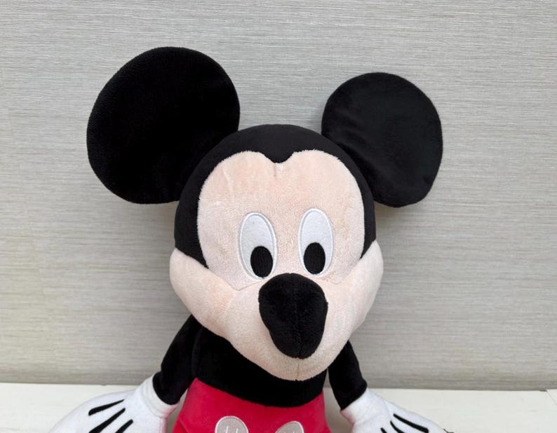 May include: A plush Mickey Mouse toy with large black ears, a black nose, and white gloves. The toy has a red pair of shorts and a cream-colored face. The toy is a classic Disney character.