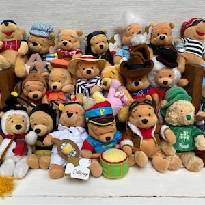Puede incluir: Una colección de peluches de Winnie the Pooh, cada uno vestido con un atuendo único. Los peluches presentan varios sombreros, disfraces y accesorios, incluyendo un gorro de marinero, un sombrero de vaquero y una bandana de pirata. Los juguetes están colocados en una estante de madera.