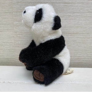 May include: A plush panda bear toy, featuring a black and white color scheme. The panda has black patches around its eyes, ears, and limbs, with a white body. The toy is sitting with its arms and legs bent, and has brown paws.