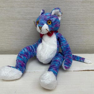 May include: A handmade plush cat toy with blue and purple striped fabric. The cat has white accents on its chest and paws, a red bow tie, and yellow eyes. The toy is sitting on a wooden surface.