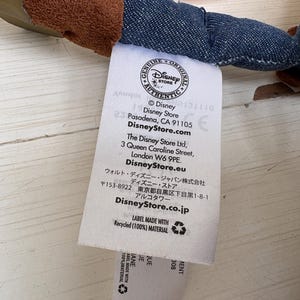 May include: A white label with text from the Disney Store, including the Disney Store logo, address, and website. The label also states it is made with recycled material.