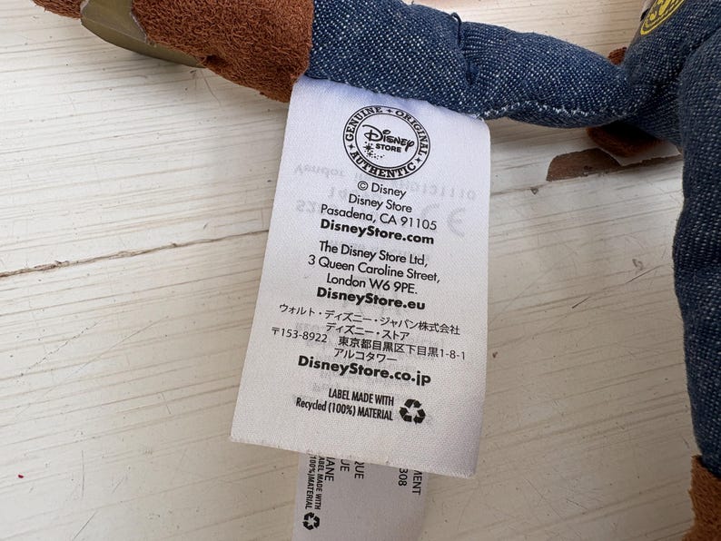 May include: A white label with text from the Disney Store, including the Disney Store logo, address, and website. The label also states it is made with recycled material.
