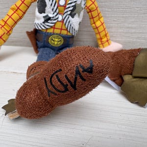 May include: A plush toy of a cowboy character, featuring a yellow plaid shirt, blue jeans, and a brown vest. The character's brown boot has the name "ANDY" written on it in black. The toy is a children's collectible.