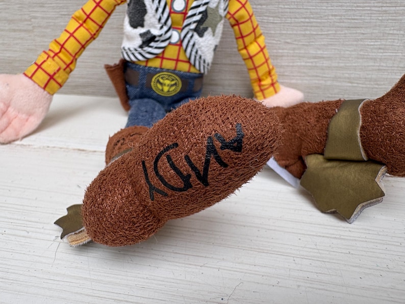 May include: A plush toy of a cowboy character, featuring a yellow plaid shirt, blue jeans, and a brown vest. The character's brown boot has the name "ANDY" written on it in black. The toy is a children's collectible.