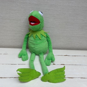 Könnte beinhalten: Ein hellgrünes Frosch-Stofftier mit einem weit geöffneten roten Mund, weiß-schwarzen Augen und einem spitzen Kragen. Die Figur hat lange, dünne Arme und Beine mit Schwimmfüßen und besteht aus weichem Stoffmaterial in sitzender Position.