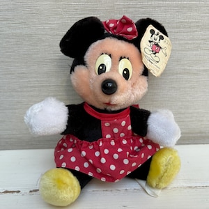 K&ouml;nnte beinhalten: Ein Pl&uuml;sch Minnie Mouse-Puppe mit einem roten Polka-Dot-Kleid, einer passenden Schleife und gelben Schuhen. Die Puppe hat schwarze Ohren und ein angebrachtes Etikett. Die Puppe sitzt auf einer wei&szlig;en Oberfl&auml;che.