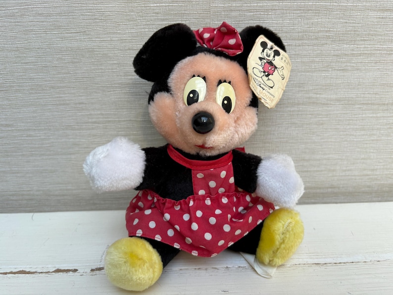 K&ouml;nnte beinhalten: Ein Pl&uuml;sch Minnie Mouse-Puppe mit einem roten Polka-Dot-Kleid, einer passenden Schleife und gelben Schuhen. Die Puppe hat schwarze Ohren und ein angebrachtes Etikett. Die Puppe sitzt auf einer wei&szlig;en Oberfl&auml;che.