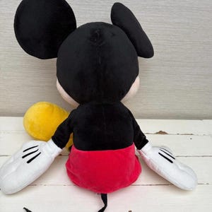 May include: A plush Mickey Mouse toy, viewed from the back. The toy features a black head and ears, a black body, red shorts, white gloves with black lines, and a long black tail. A yellow object is visible.