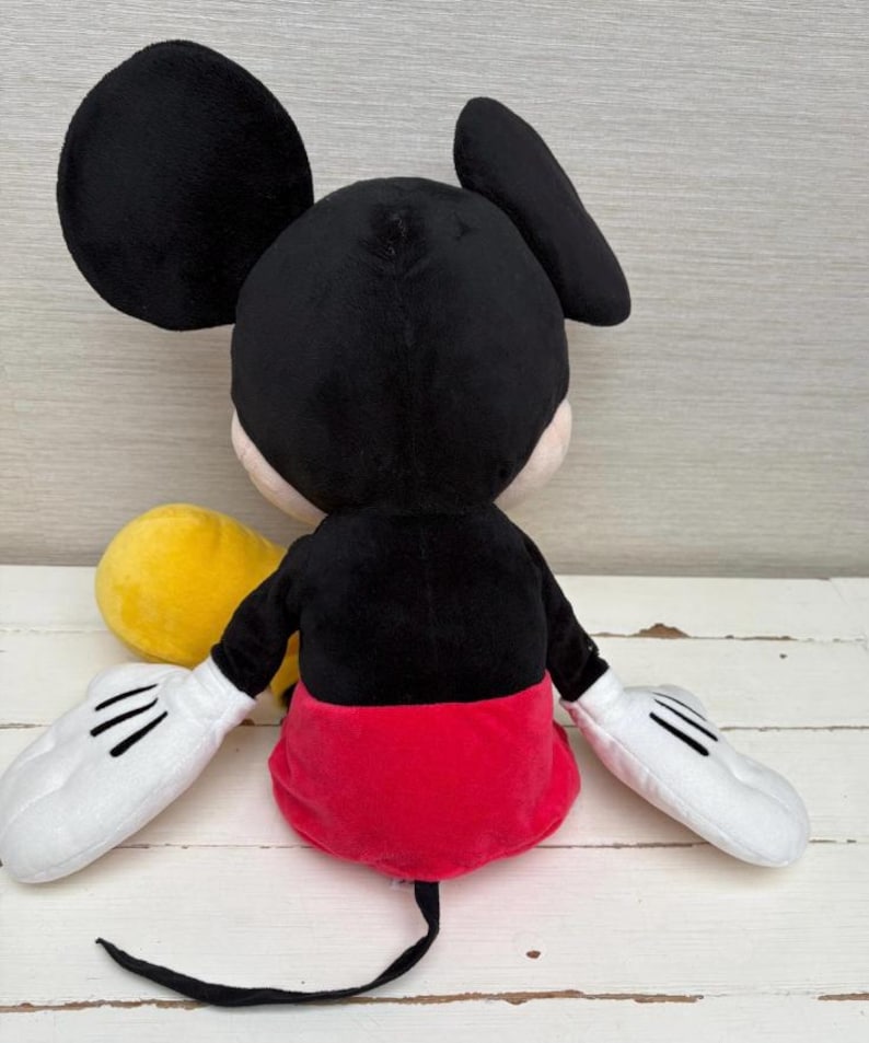 May include: A plush Mickey Mouse toy, viewed from the back. The toy features a black head and ears, a black body, red shorts, white gloves with black lines, and a long black tail. A yellow object is visible.