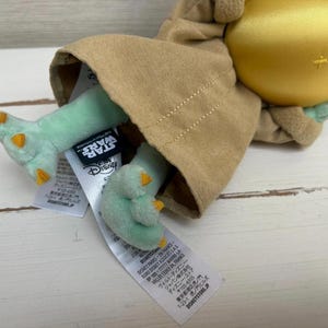 Pode incluir: Um brinquedo de pel&uacute;cia de um personagem da franquia Star Wars. O brinquedo tem uma cabe&ccedil;a grande, p&eacute;s verdes com garras laranja e est&aacute; vestindo uma t&uacute;nica marrom. Os logotipos da Star Wars e da Disney s&atilde;o vis&iacute;veis nas etiquetas anexadas.