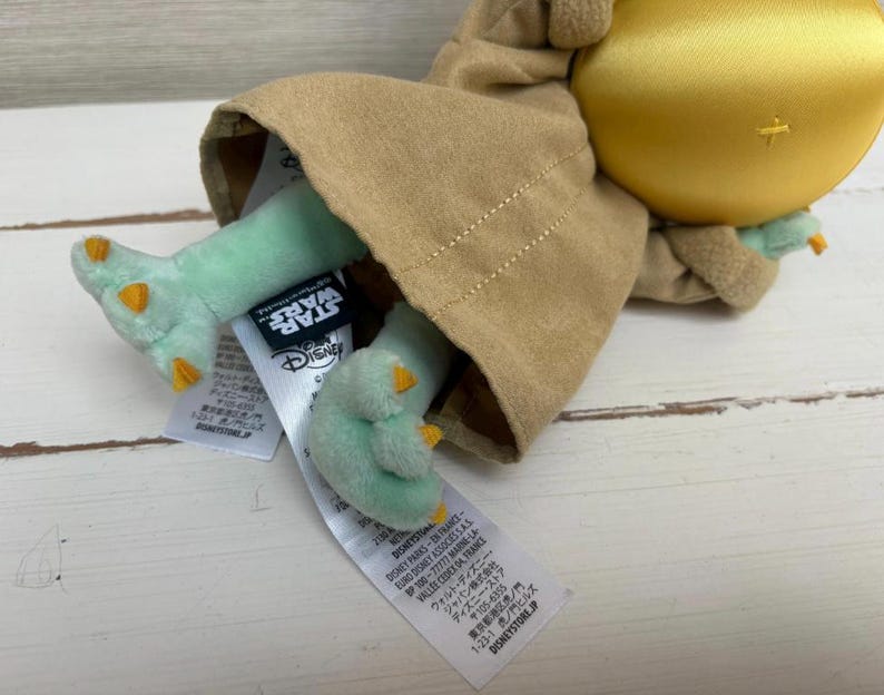 Pode incluir: Um brinquedo de pel&uacute;cia de um personagem da franquia Star Wars. O brinquedo tem uma cabe&ccedil;a grande, p&eacute;s verdes com garras laranja e est&aacute; vestindo uma t&uacute;nica marrom. Os logotipos da Star Wars e da Disney s&atilde;o vis&iacute;veis nas etiquetas anexadas.