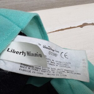 Może przedstawiać: Zbliżenie pluszowej zabawki z turkusową tkaniną i metką z napisem "Liberty Minnie" i "&copy;DISNEY". Etykieta zawiera informacje o produkcji. Zabawka jest wyprodukowana w Chinach.