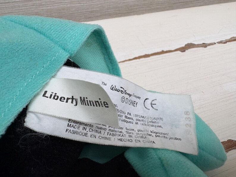 Może przedstawiać: Zbliżenie pluszowej zabawki z turkusową tkaniną i metką z napisem "Liberty Minnie" i "&copy;DISNEY". Etykieta zawiera informacje o produkcji. Zabawka jest wyprodukowana w Chinach.